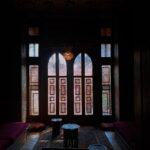 صميم داخلي لغرفة تراثية مصرية يبرز فيها نافذة مشربية خشبية ضخمة يتسلل منها ضوء الشمس ليرسم ظلالاً هندسية على سجادة شرقية يدوية ووسائد أرضية تقليدية.