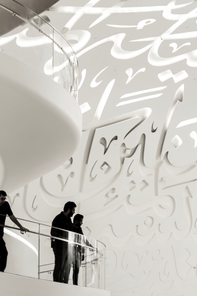 تصميم معماري داخلي مودرن باللون الأبيض يبرز فيه حائط وسقف ممتد منقوش بحروف الخط العربي البارزة والمفرغة للإضاءة، مع أشخاص يقفون على شرفة زجاجية انسيابية.