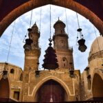 لقطة من داخل إيوان مدرسة مملوكية قديمة تطل عبر عقد حجري ضخم على صحن مكشوف، وتظهر في الخلفية مئذنتان حجريتان أثريتان وقبة مع قناديل ثريا نحاسية متدلية.