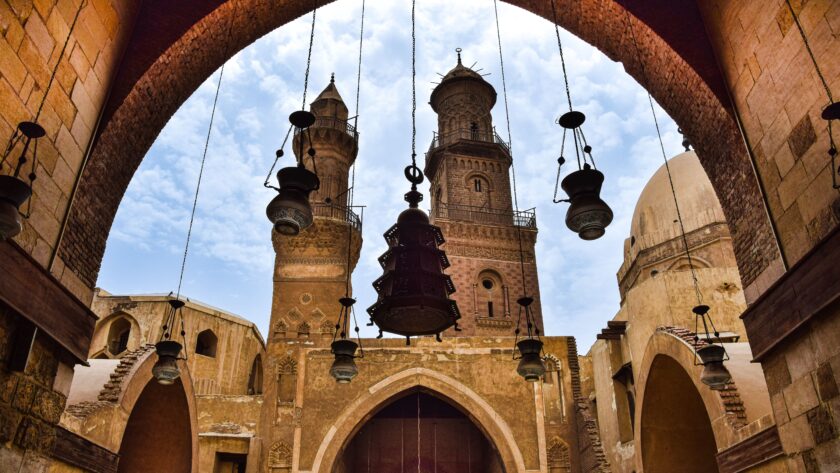 لقطة من داخل إيوان مدرسة مملوكية قديمة تطل عبر عقد حجري ضخم على صحن مكشوف، وتظهر في الخلفية مئذنتان حجريتان أثريتان وقبة مع قناديل ثريا نحاسية متدلية.