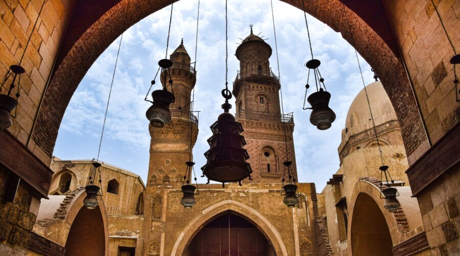 لقطة من داخل إيوان مدرسة مملوكية قديمة تطل عبر عقد حجري ضخم على صحن مكشوف، وتظهر في الخلفية مئذنتان حجريتان أثريتان وقبة مع قناديل ثريا نحاسية متدلية.