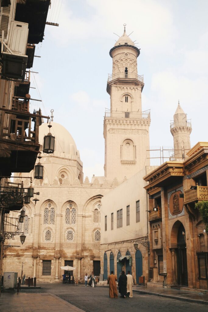 مشهد شارع تاريخي ضيق في القاهرة القديمة، يُظهر واجهات المساجد والمباني الحجرية الأثرية من العصر المملوكي، وتبرز مصطلحات العمارة الإسلامية بوضوح مثل المئذنة الأسطوانية الطويلة والقبة المستديرة ونوافذ المشربية، بينما يسير مارة بملابس تقليدية في الزقاق