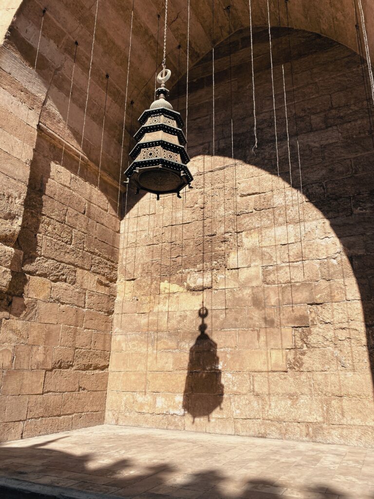 ثريا نحاسية تراثية (تنورة) متدلية داخل إيوان مسجد أثري، ويلوح ظلها على الجدار الحجري ليبرز مصطلحات العمارة الإسلامية وتناغم الضوء والظل في التصميم القديم